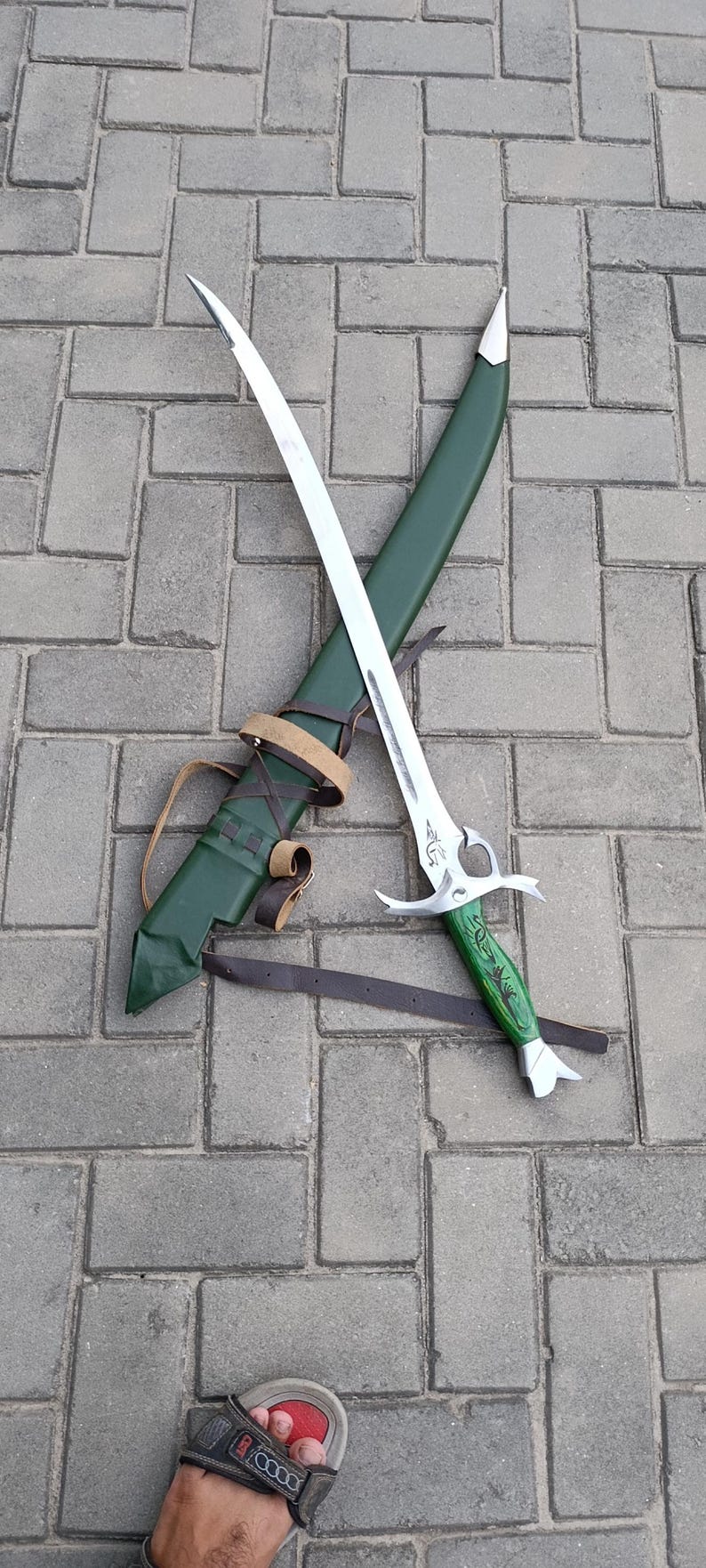 Puede incluir: Una espada plateada con una hoja curva y un mango verde, apoyada sobre una vaina verde. La espada tiene un dise&ntilde;o ornamentado en el mango y una correa de cuero marr&oacute;n.