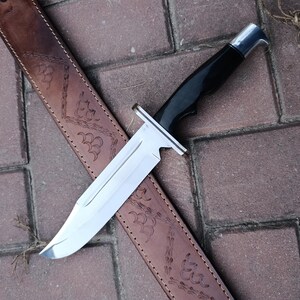 May include: A large hunting knife with a polished steel blade and a black handle rests on a brown leather sheath. The sheath is decorated with an embossed floral pattern. The knife is designed for outdoor use.