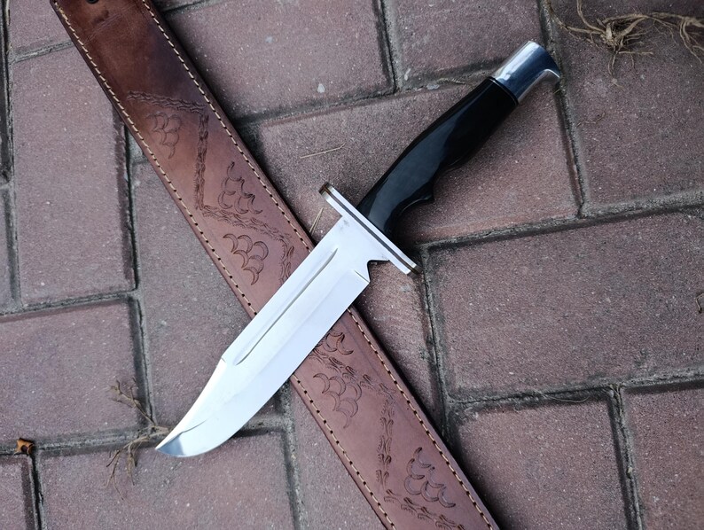 May include: A large hunting knife with a polished steel blade and a black handle rests on a brown leather sheath. The sheath is decorated with an embossed floral pattern. The knife is designed for outdoor use.