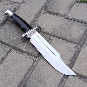 May include: A large, polished hunting knife with a shiny, silver blade and a black handle. The knife has a silver bolster and a curved blade. The handle is smooth and appears to be made of a durable material.