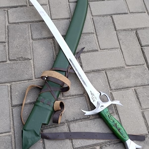 Puede incluir: Una espada de fantas&iacute;a plateada con una hoja curva y un mango verde, apoyada sobre una vaina verde con correas de cuero marr&oacute;n. La espada tiene grabados decorativos y una empu&ntilde;adura plateada.
