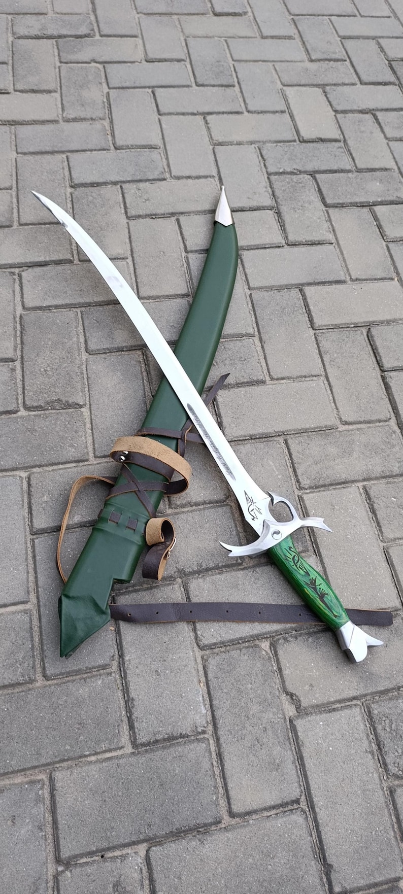 Puede incluir: Una espada de fantas&iacute;a plateada con una hoja curva y un mango verde, apoyada sobre una vaina verde con correas de cuero marr&oacute;n. La espada tiene grabados decorativos y una empu&ntilde;adura plateada.
