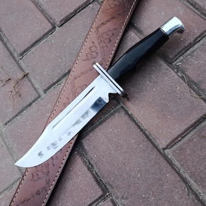 May include: A hunting knife with a shiny, silver blade and a black handle rests on a brown leather sheath. The knife has a silver bolster and a curved handle. The sheath is embossed with a decorative pattern.