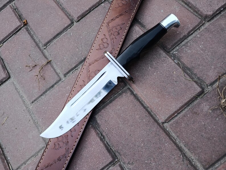 May include: A hunting knife with a shiny, silver blade and a black handle rests on a brown leather sheath. The knife has a silver bolster and a curved handle. The sheath is embossed with a decorative pattern.