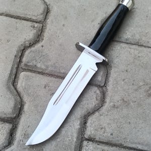 May include: A large, polished stainless steel knife with a black handle and silver accents. The blade has a sharp point and a double groove design. The knife is designed for outdoor use and is a hunting or survival tool.