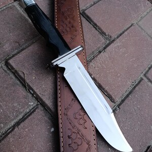 May include: A hunting knife with a polished steel blade and a black handle. The knife rests on a brown leather sheath with an embossed design. The handle has a silver bolster.