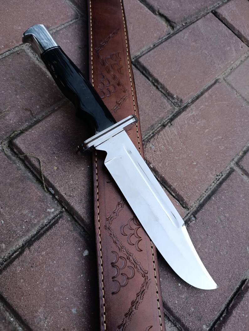 May include: A hunting knife with a polished steel blade and a black handle. The knife rests on a brown leather sheath with an embossed design. The handle has a silver bolster.