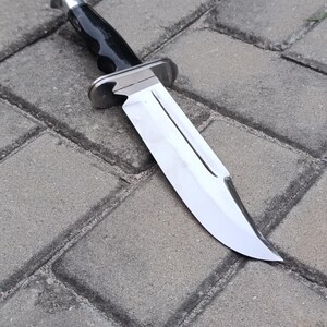 May include: A large, stainless steel knife with a polished blade and a black handle. The knife features a silver bolster and a curved blade, designed for cutting. The knife is set against a gray brick background.