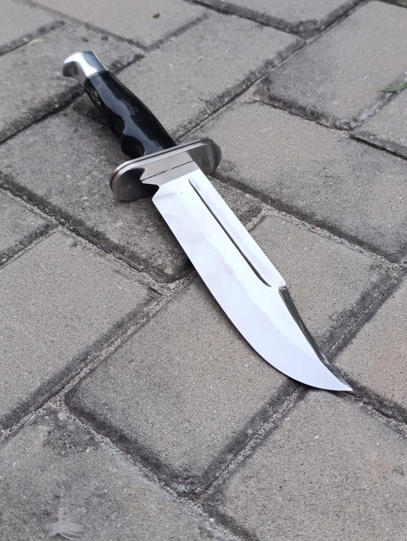 May include: A large, stainless steel knife with a polished blade and a black handle. The knife features a silver bolster and a curved blade, designed for cutting. The knife is set against a gray brick background.
