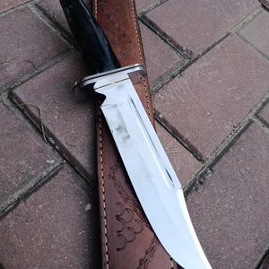 May include: A large hunting knife with a polished steel blade and a black handle. The knife rests on a brown leather sheath with decorative embossed patterns. The blade is approximately 8 inches long.