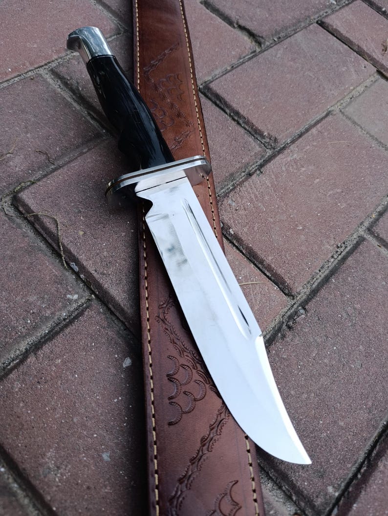May include: A large hunting knife with a polished steel blade and a black handle. The knife rests on a brown leather sheath with decorative embossed patterns. The blade is approximately 8 inches long.
