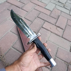May include: A large hunting knife with a polished steel blade and a black handle. The knife has a silver bolster and a silver end cap. The knife is shown with a brown leather sheath.