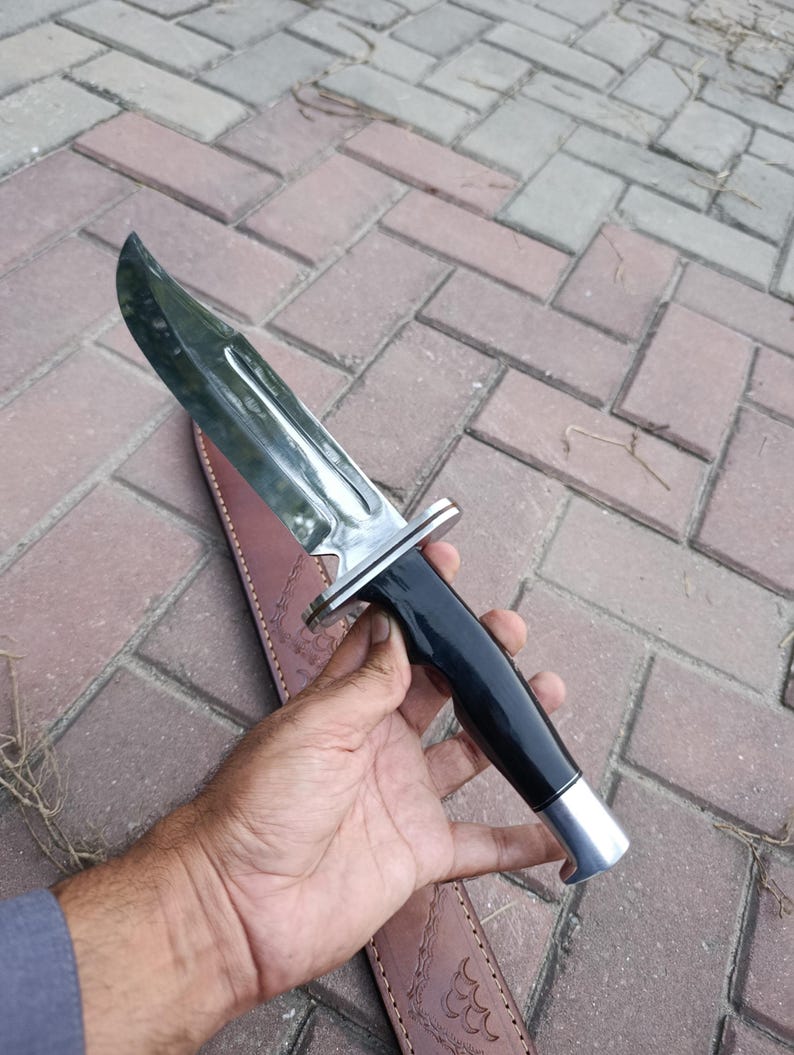 May include: A large hunting knife with a polished steel blade and a black handle. The knife has a silver bolster and a silver end cap. The knife is shown with a brown leather sheath.