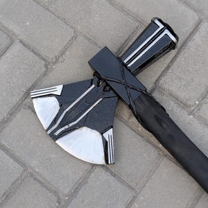 May include: A black and silver axe with a long black handle. The axe head has a curved blade with silver accents and a black body. The handle is wrapped in a dark material. The axe is lying on a gray brick surface.