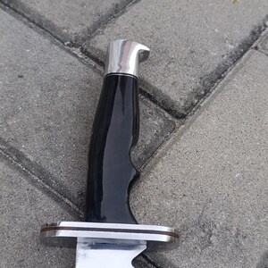 May include: Close-up of a large knife with a shiny, silver blade and a black handle. The handle has a curved design for grip and is topped with a silver cap. The knife is set against a gray, textured background.