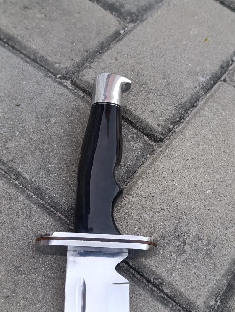 May include: Close-up of a large knife with a shiny, silver blade and a black handle. The handle has a curved design for grip and is topped with a silver cap. The knife is set against a gray, textured background.