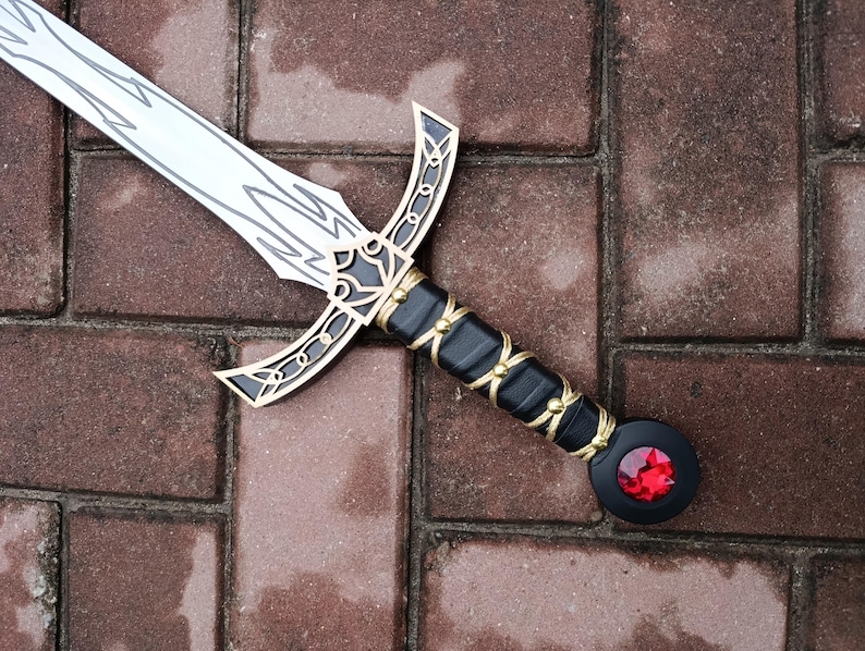 May include: A decorative sword with a silver blade and intricate gold and black detailing. The hilt is black with gold accents and a red gem at the pommel. The sword is lying on a brick surface.