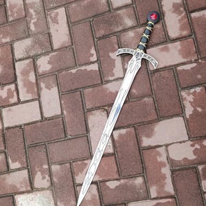 May include: A silver sword with a decorative hilt featuring black and gold accents, a red gem, and a detailed blade design. The sword is lying on a brick surface.