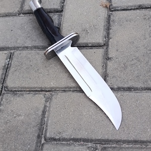 May include: A large, stainless steel knife with a black handle and a silver bolster. The blade is wide and has a curved edge. The knife is lying on a gray brick surface.