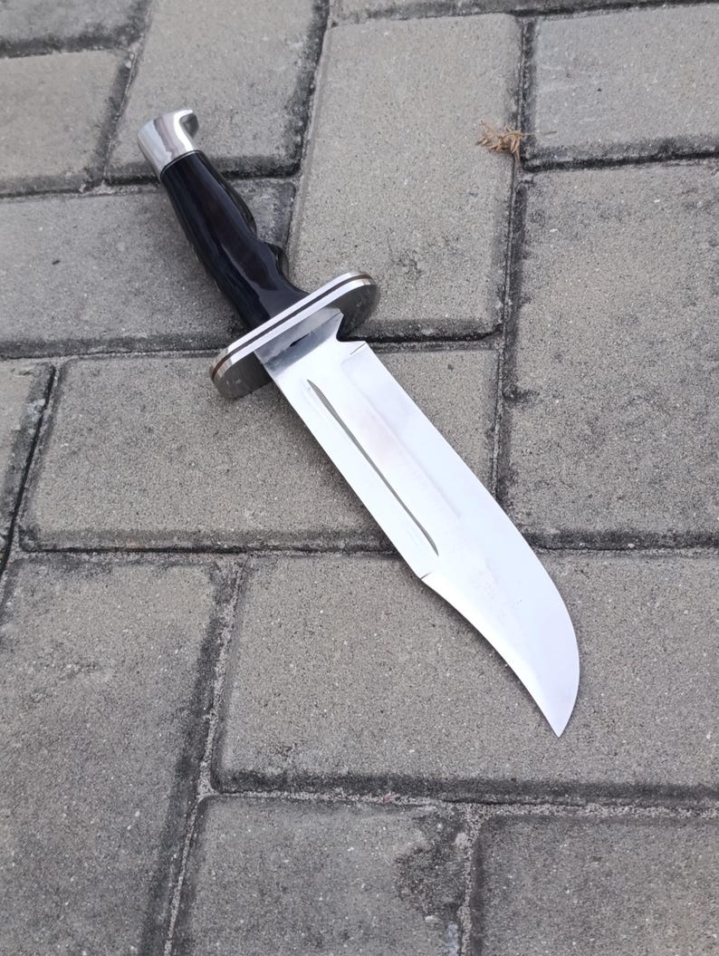 May include: A large, stainless steel knife with a black handle and a silver bolster. The blade is wide and has a curved edge. The knife is lying on a gray brick surface.