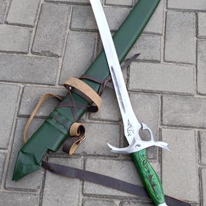 Puede incluir: Una espada de fantas&iacute;a plateada y verde con una vaina verde a juego y correas de cuero marr&oacute;n. La espada tiene una hoja curva y un mango verde detallado. La vaina es verde y tiene una correa de cuero para llevar.