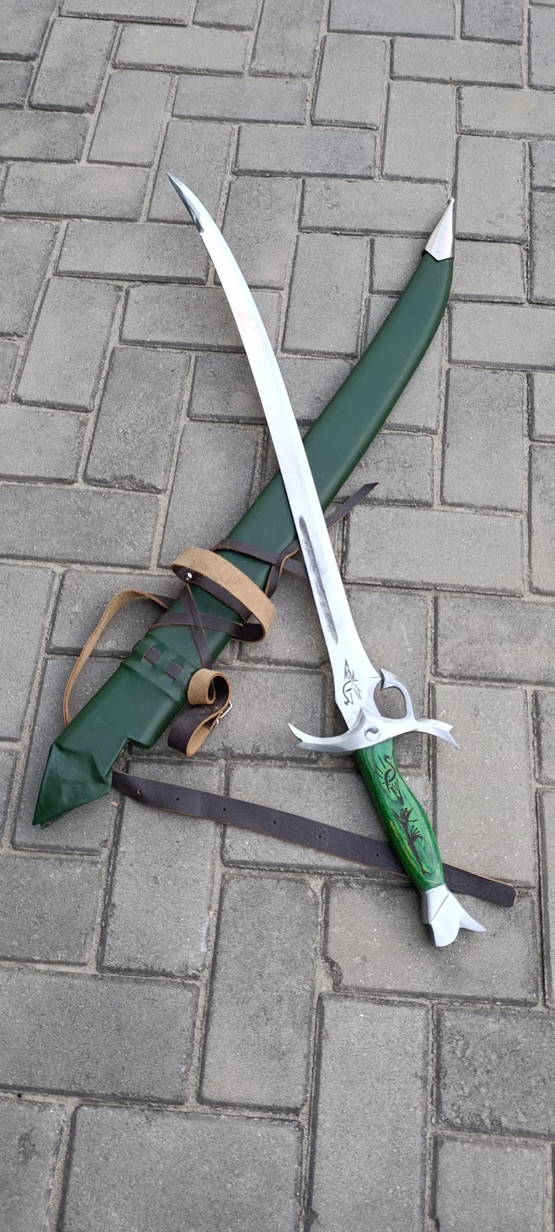 Puede incluir: Una espada de fantas&iacute;a plateada y verde con una vaina verde a juego y correas de cuero marr&oacute;n. La espada tiene una hoja curva y un mango verde detallado. La vaina es verde y tiene una correa de cuero para llevar.