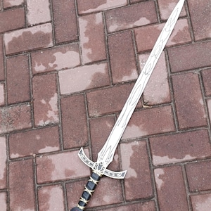 May include: A silver-colored sword with a decorative blade and a detailed hilt. The hilt features black and gold accents, with a red gem at the pommel. The sword is lying on a brick surface.