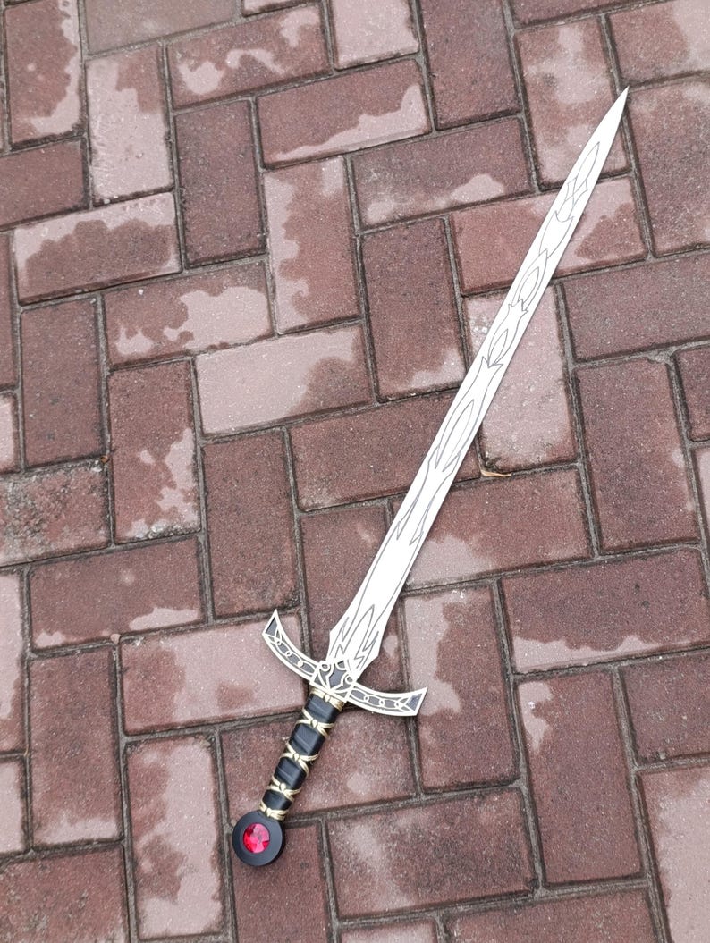 May include: A silver-colored sword with a decorative blade and a detailed hilt. The hilt features black and gold accents, with a red gem at the pommel. The sword is lying on a brick surface.