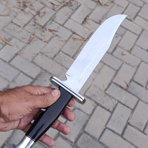 May include: A large, polished hunting knife with a curved blade. The knife features a black handle with silver accents at the base and guard. The blade is bright and reflective, and the knife is held against a neutral background.