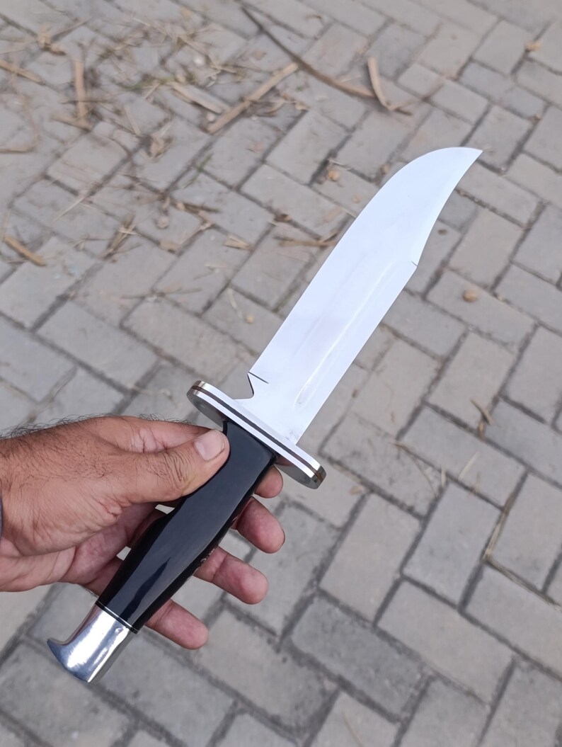 May include: A large, polished hunting knife with a curved blade. The knife features a black handle with silver accents at the base and guard. The blade is bright and reflective, and the knife is held against a neutral background.