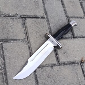 May include: A large hunting knife with a polished, silver blade and a black handle. The knife features a silver bolster and a curved blade, designed for outdoor use and survival.