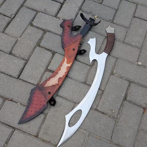 Könnte beinhalten: Ein geschwungenes Metallschwert mit silberner Klinge, einer Aussparung nahe der Spitze und einem braun umwickelten Griff, neben einer braunen Lederscheide mit dunkelrotem Farbverlauf, Rautenmuster und schwarzen Metalldruckknöpfen.