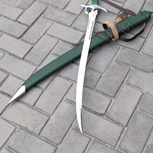 Puede incluir: Una espada curva con una hoja plateada y un mango verde descansa junto a su vaina verde. La vaina tiene una punta plateada y est&aacute; asegurada con una correa de cuero marr&oacute;n. La espada est&aacute; sobre una superficie de ladrillos grises.