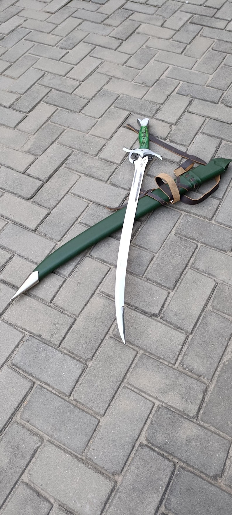 Puede incluir: Una espada curva con una hoja plateada y un mango verde descansa junto a su vaina verde. La vaina tiene una punta plateada y est&aacute; asegurada con una correa de cuero marr&oacute;n. La espada est&aacute; sobre una superficie de ladrillos grises.