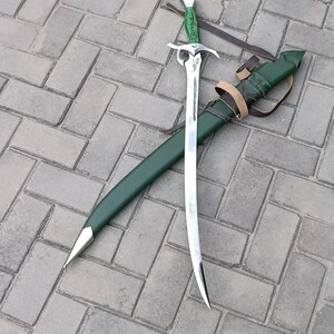 Puede incluir: Una espada plateada con una hoja curva y un mango verde descansa sobre una vaina verde. La espada tiene una empu&ntilde;adura decorativa y est&aacute; acompa&ntilde;ada de una correa de cuero. Los objetos est&aacute;n sobre una superficie de ladrillo gris.