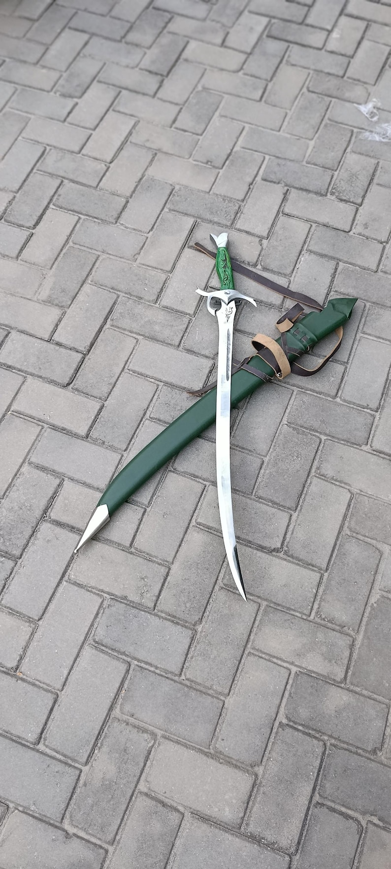 Puede incluir: Una espada plateada con una hoja curva y un mango verde descansa sobre una vaina verde. La espada tiene una empu&ntilde;adura decorativa y est&aacute; acompa&ntilde;ada de una correa de cuero. Los objetos est&aacute;n sobre una superficie de ladrillo gris.
