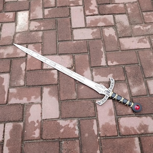 May include: A silver-colored sword with a decorative handle featuring black and gold accents and a red gem. The sword's blade has a detailed design. The sword is lying on a brick surface.