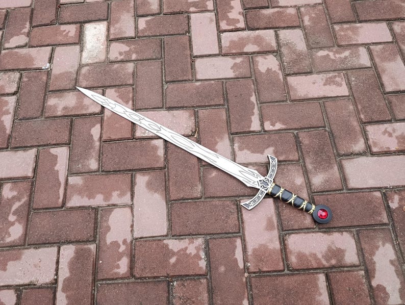May include: A silver-colored sword with a decorative handle featuring black and gold accents and a red gem. The sword's blade has a detailed design. The sword is lying on a brick surface.