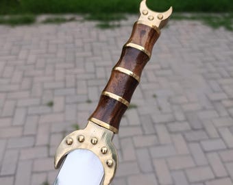 May include: A close-up of a decorative sword with a polished silver blade. The handle is made of dark wood with brass accents and a crescent-shaped pommel. The sword is likely for display or ceremonial purposes.