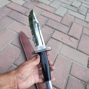 May include: A large, polished steel knife with a black handle and a silver bolster. The knife has a curved blade and is accompanied by a brown leather sheath. The knife is designed for outdoor use.
