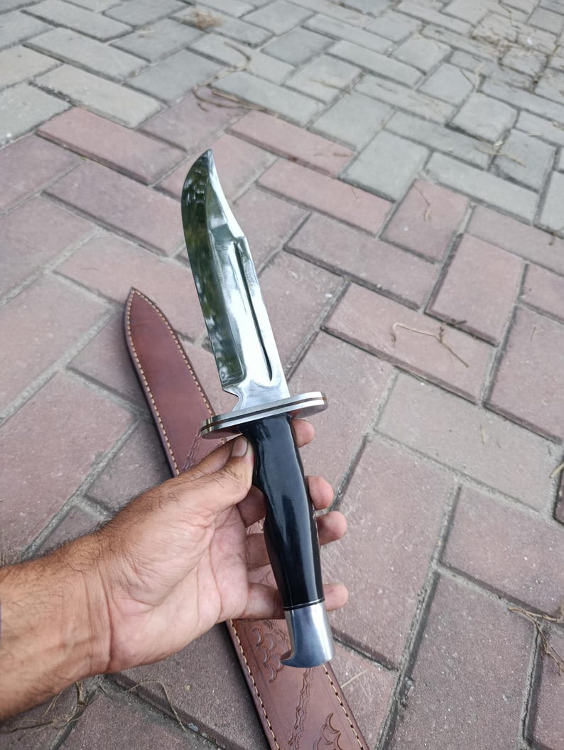 May include: A large, polished steel knife with a black handle and a silver bolster. The knife has a curved blade and is accompanied by a brown leather sheath. The knife is designed for outdoor use.