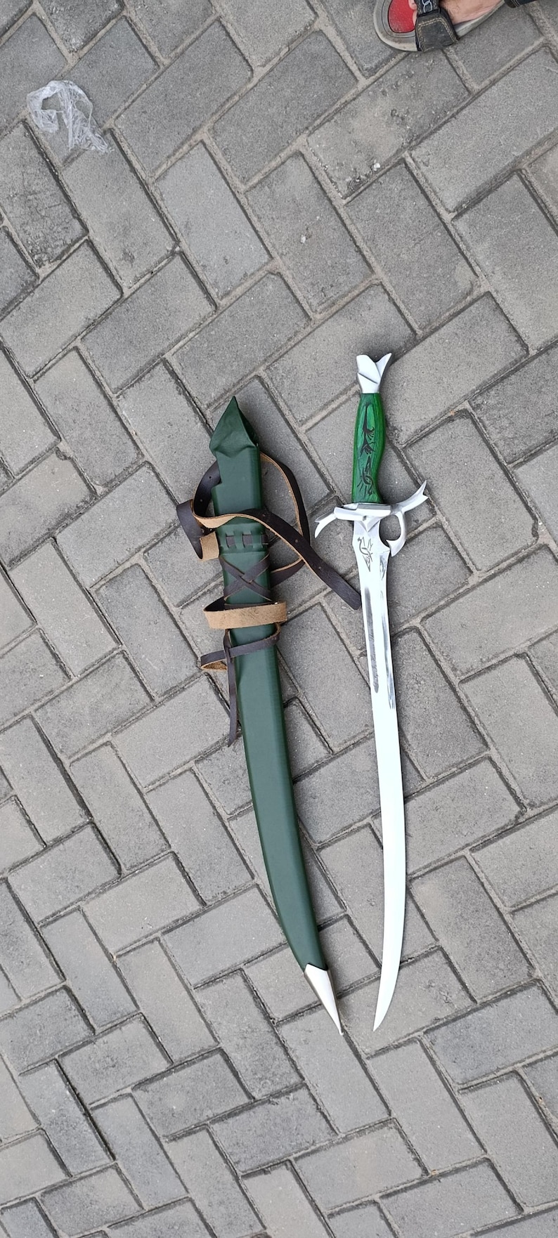 Puede incluir: Una espada de fantas&iacute;a verde y plateada con una hoja curva, junto a una vaina verde a juego. La espada tiene una empu&ntilde;adura verde decorativa y una guarda plateada. La vaina est&aacute; asegurada con correas de cuero marr&oacute;n.