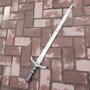 May include: A silver sword with a decorative hilt and a red gem at the base of the handle. The blade has etched designs. The handle is black and gold. The sword is lying on a brick surface.