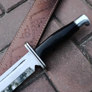 May include: Close-up of a hunting knife with a shiny, silver blade and a black handle. The knife is partially sheathed in a brown leather case with decorative embossed patterns. The knife's blade is approximately 8 inches long.