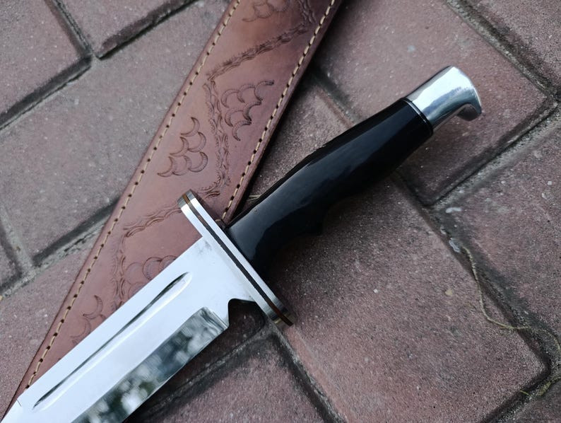 May include: Close-up of a hunting knife with a shiny, silver blade and a black handle. The knife is partially sheathed in a brown leather case with decorative embossed patterns. The knife's blade is approximately 8 inches long.