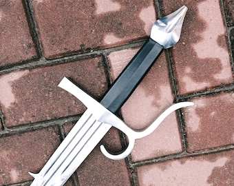 May include: A silver sword with a black handle and a distinctive crossguard. The blade has a series of parallel grooves. The sword is resting on a brick surface.
