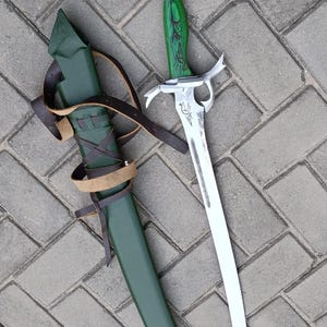 Puede incluir: Una espada decorativa con una hoja curva y un mango verde con tallas ornamentadas. La espada est&aacute; junto a una vaina verde con correas de cuero marr&oacute;n. La hoja de la espada es plateada y el mango es verde.