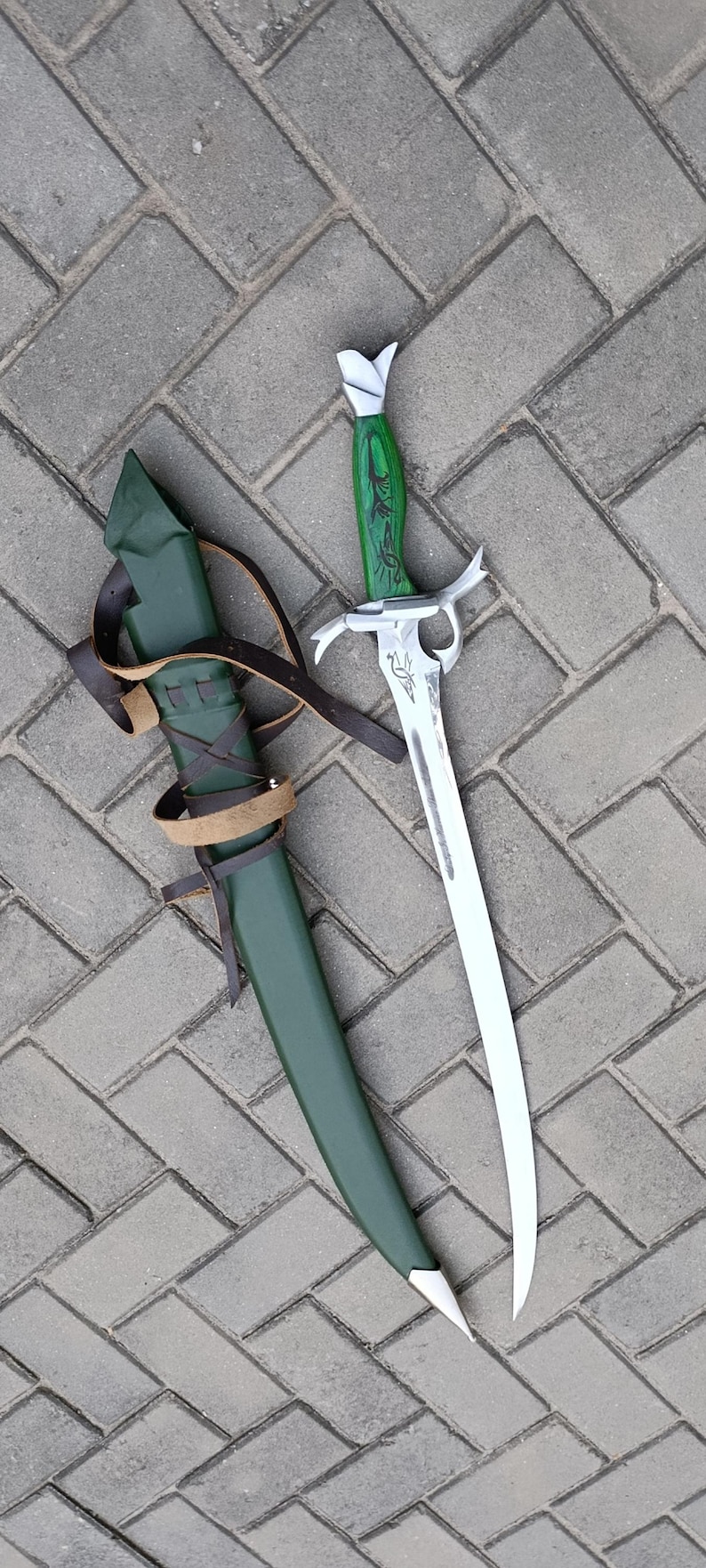 Puede incluir: Una espada decorativa con una hoja curva y un mango verde con tallas ornamentadas. La espada est&aacute; junto a una vaina verde con correas de cuero marr&oacute;n. La hoja de la espada es plateada y el mango es verde.