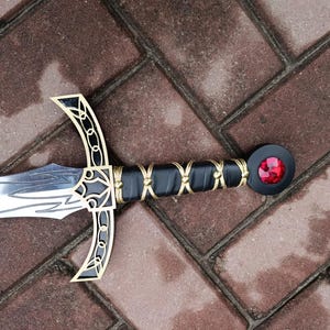 May include: A decorative sword with a silver blade and a black handle wrapped with gold-colored wire. The crossguard and pommel feature gold and black accents, and the pommel has a red jewel. The sword is lying on a brick surface.
