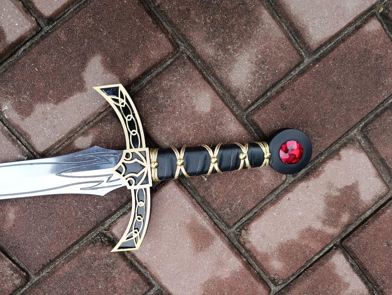 May include: A decorative sword with a silver blade and a black handle wrapped with gold-colored wire. The crossguard and pommel feature gold and black accents, and the pommel has a red jewel. The sword is lying on a brick surface.
